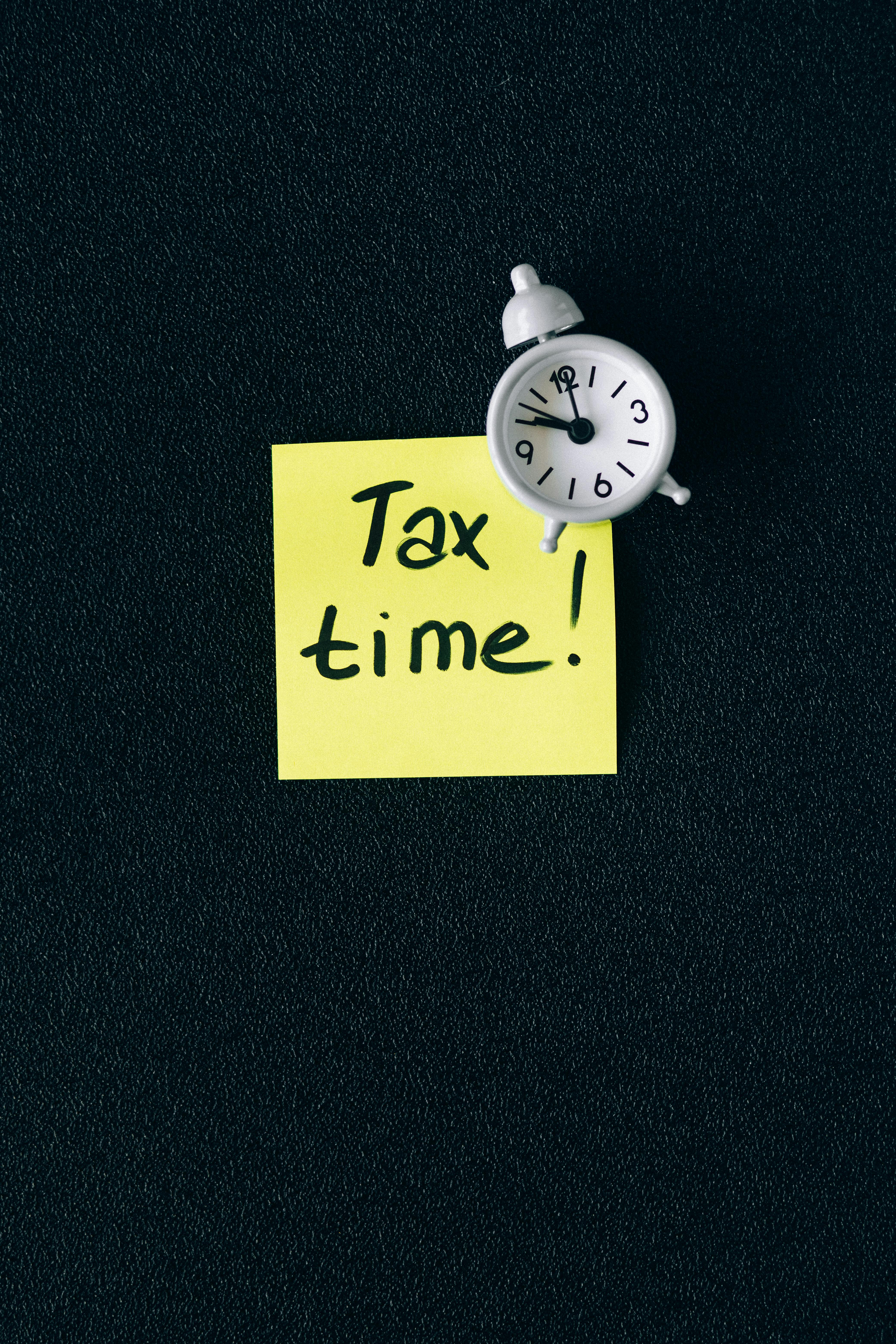 Black background with a yellow post-it note and the words "Tax time!" written in black texta and a small white alarm clock placed over the right hand top corner of the post-it note