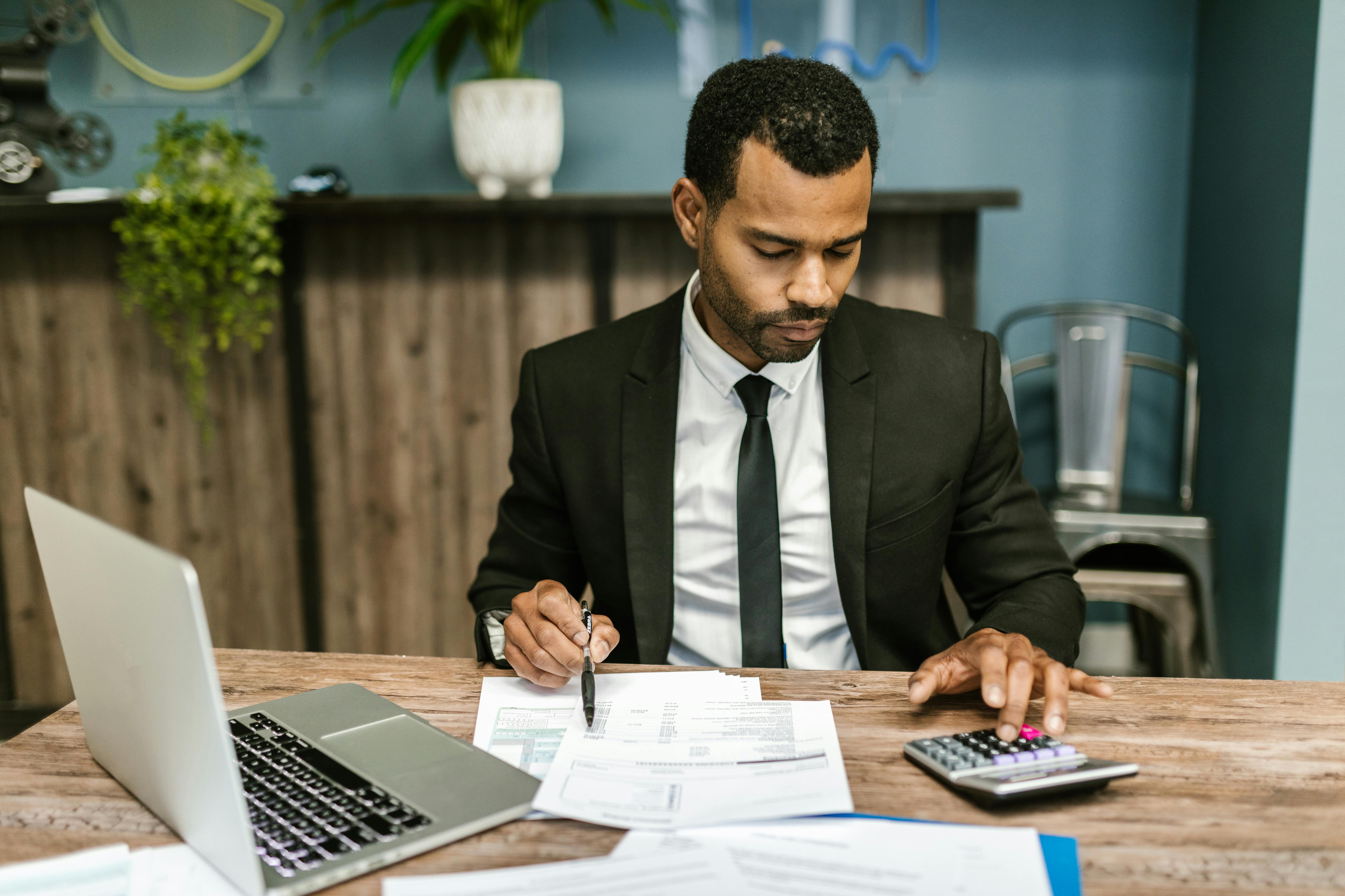 An accountant working at a desk using a calculator with paperwork and an open laptop on the desk